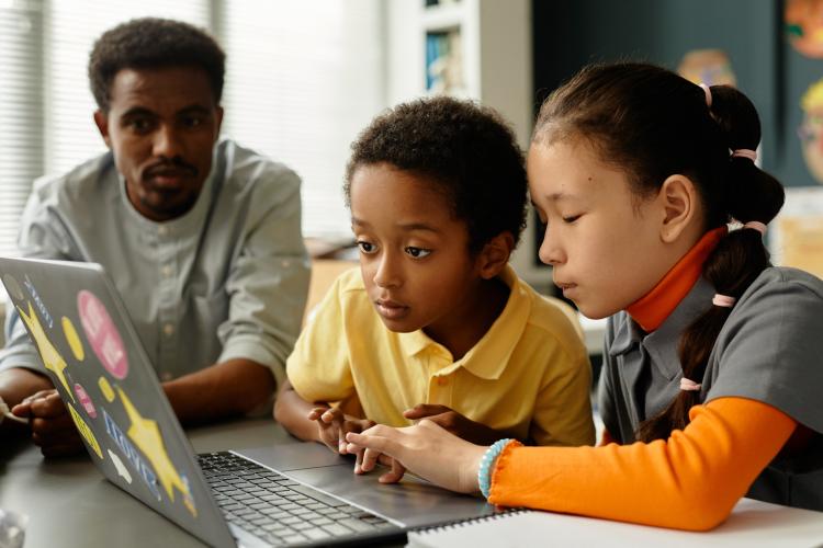 Two children intently using laptop with teacher nearby, supervising