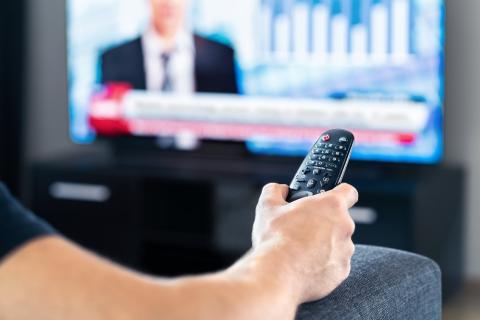 Person holds tv remote with news on tv blurred out in background