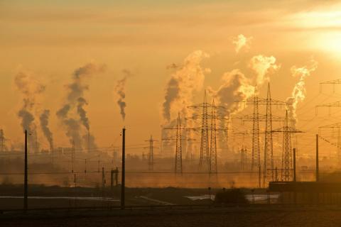 Smoke and steam rise from numerous industrial chimneys at sunset, surrounded by power lines and transmission towers, illustrating heavy air pollution. 