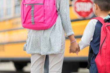 Sister holding brothers hand, backpacks on their backs, with blurred school bus in background
