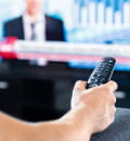 Person's hand holding a remote in the foreground with news channel blurred out on tv in background