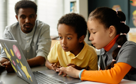 Two children intently using laptop with teacher nearby, supervising