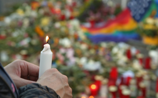 Person's hand holding lit candle at a vigil with blurred candles, flowers, and pride flag